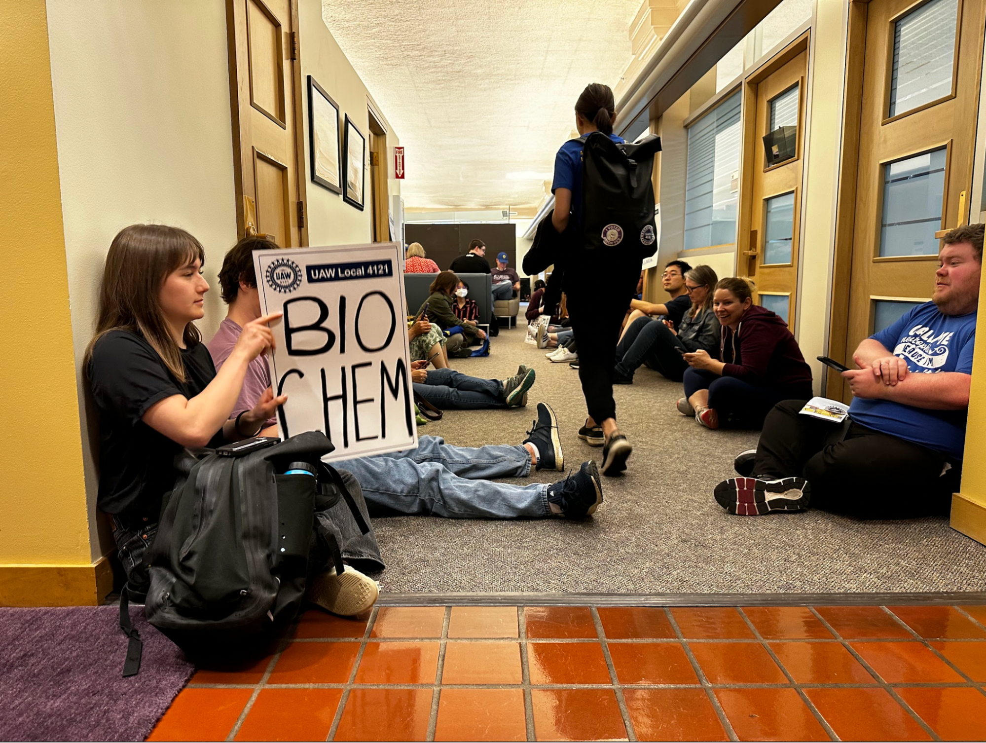 Postdocs, RSEs, and supporters sit on the floor in a hallway. One person with union stickers on their backpack is walking past the camera. Another person in the forground holds a UAW sign that reads "BIO-CHEM"