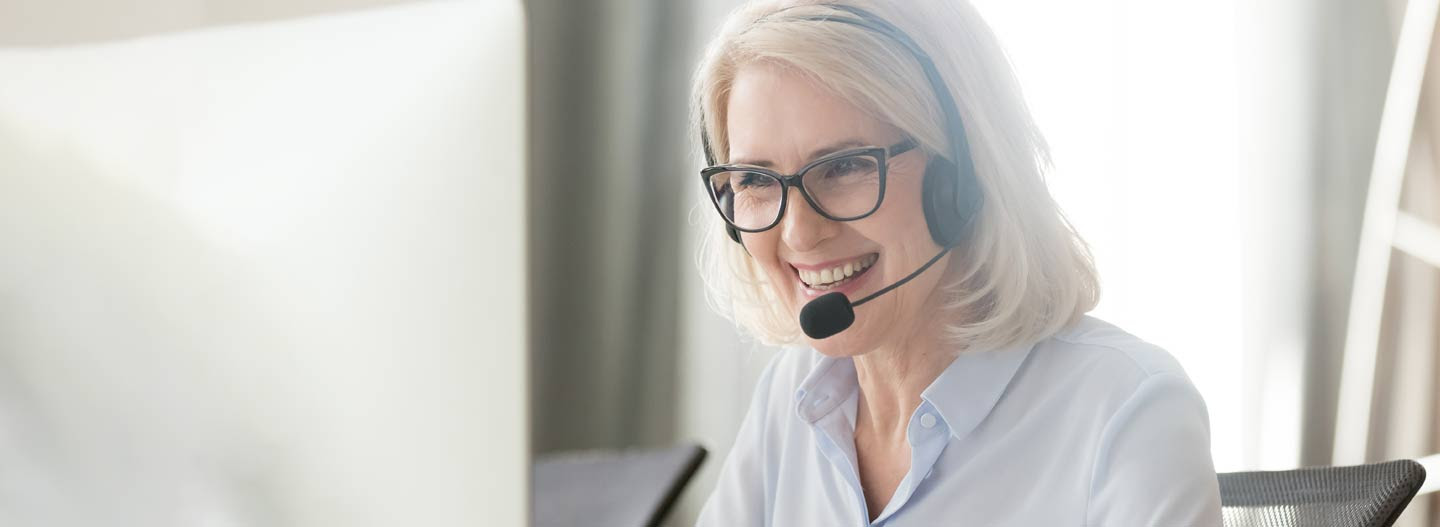 Smiling senior woman wearing headset sitting in front of a computer monitor.