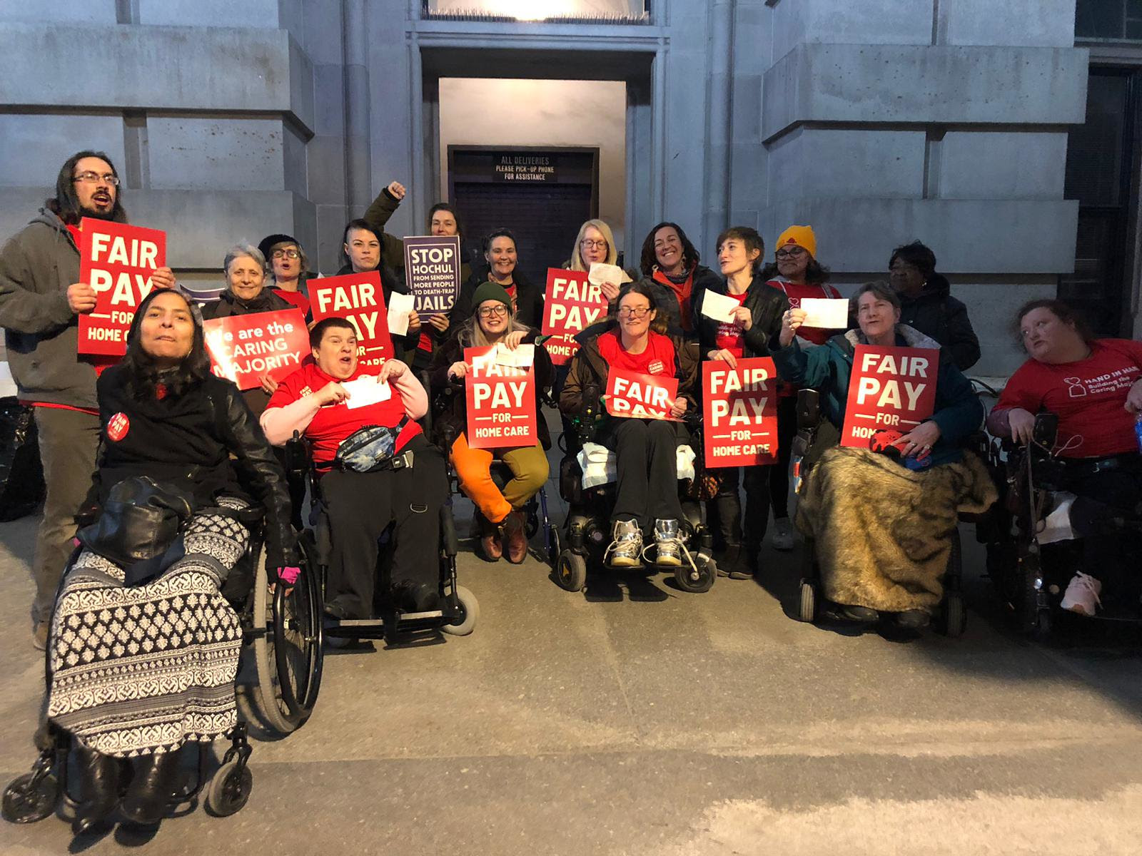 Photo of a group of people. half in wheelchairs, holding fair pay for home care signs
