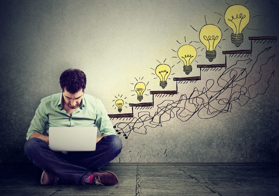 A man sits on the floor with his laptop; on the wall behind him is a drawing of stairs. Each stair has a lightbulb that is bigger than the last. A man sits on the floor with his laptop; on the wall behind him is a drawing of stairs. Each stair has a lightbulb that is bigger than the last.