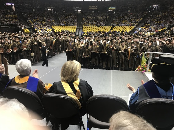 University of Wyoming graduates, in the caps and gowns, fill the floor of the Arena Auditorium during their commencement ceremony while faculty on the stage applaud the graduates.