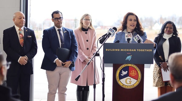 Gov. Gretchen Whitmer, at a podium with an affixed Building Michigan Together sign, flanked by two men and two women