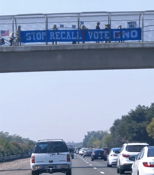 A 27-foot long banner reading "Stop Recall! Vote NO" in white text on a blue background is being carried by the Banner Brigade on a Davis freeway overpass. Heavy traffic passes on the road beneath the overpass.
