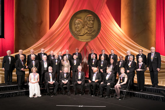 Men and women in black tie attire pose for a photo on a stage. 
