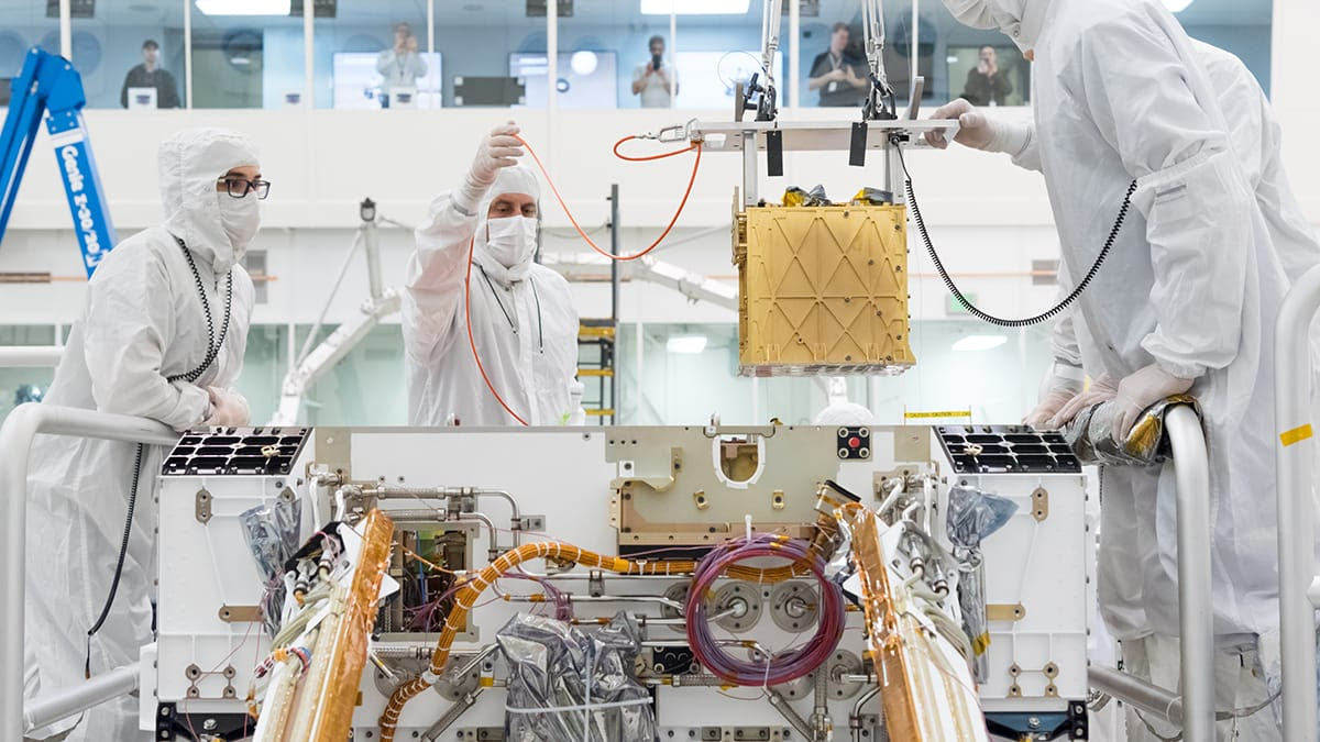 Image shows three workers in white "bunny suits“ in a clean room installing a gold, roughly cube-shaped instrument onto the chassis of the Perseverance rover, during its assembly. In the background, spectators look on from behind the glass windows of a viewing gallery.