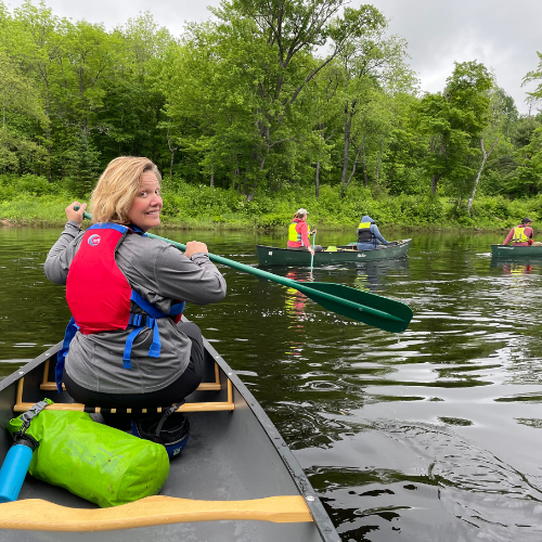A blond woman smiles at the photographer while paddling a canoe.