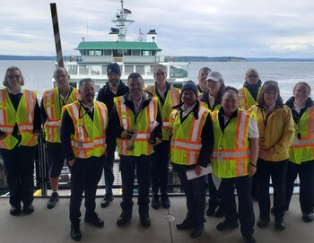 Thirteen people in safety vests posing for a photo at a ferry terminal with a vessel in the background