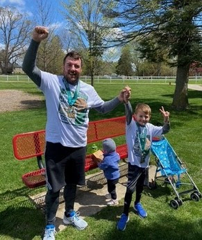 a dark-haired man dressed in running gear holds hands with a little boy, both smiling, arms raised in victory, outdoors by an orange bench