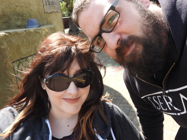 A woman and a man outdoors. The woman wears sunglasses and has long brown hair and the man wears black glasses and has a full beard