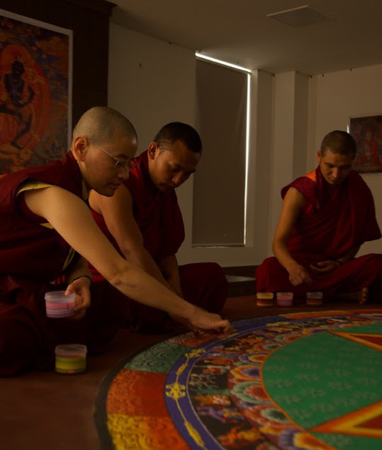 A photograph of three men making a colourful sand mandala in a dimly lit room.
