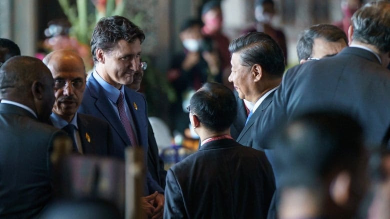 The Canadian prime minister smiles at the Chinese leader, who is accompanied by an aide. In the background, several individuals speak to each other.