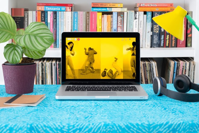 Photograph of a laptop on a desk.  On the laptop screen are four vertical photographs placed side to side, each monotone, but with a bright yellow tone overlaid. Each image shows an individual in a performative pose. On the far left a woman facing the camera just appears into view holding her arm up bent at the elbow, fingers splayed, one hand almost touching the other. The image to right of this one shows a man facing away from the camera hands pushing against a plain wall, leaning hard against it. The image to the right of this shows a man lying on his back on the floor, neck arched back with one foot raised against a plain wall. The image on the far right show a woman just disappearing out of the frame, facing away from the camera. She is standing with her legs crossed and her left arm raised level with her shoulders and bent at the elbow. On the laptop screen are videocall icons, and a red telephone icon. Around the laptop is a houseplant, lamp and headphones.
