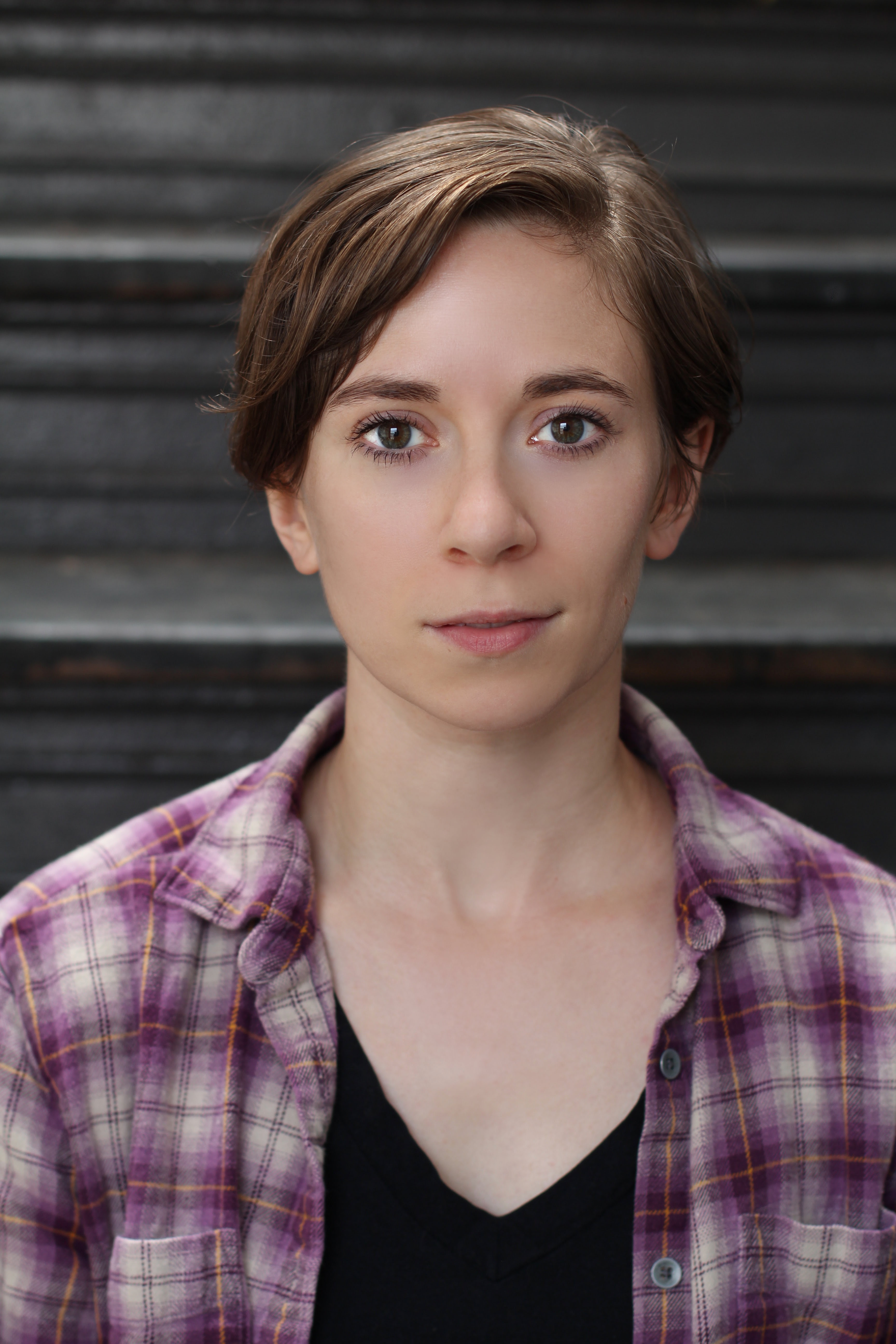 Danielle sits, looking into the camera. Her brown hair is short and parted deeply to one side. She is wearing a purple, white, and gold plaid overshirt, which is unbuttoned and shows her black v-neck tee. Her face shows the hint of a smile.