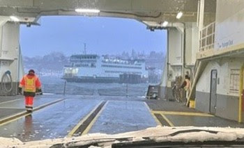 View looking out a vehicle windshield with snow on wipers, person on the car deck of ferry and another ferry off in the distance