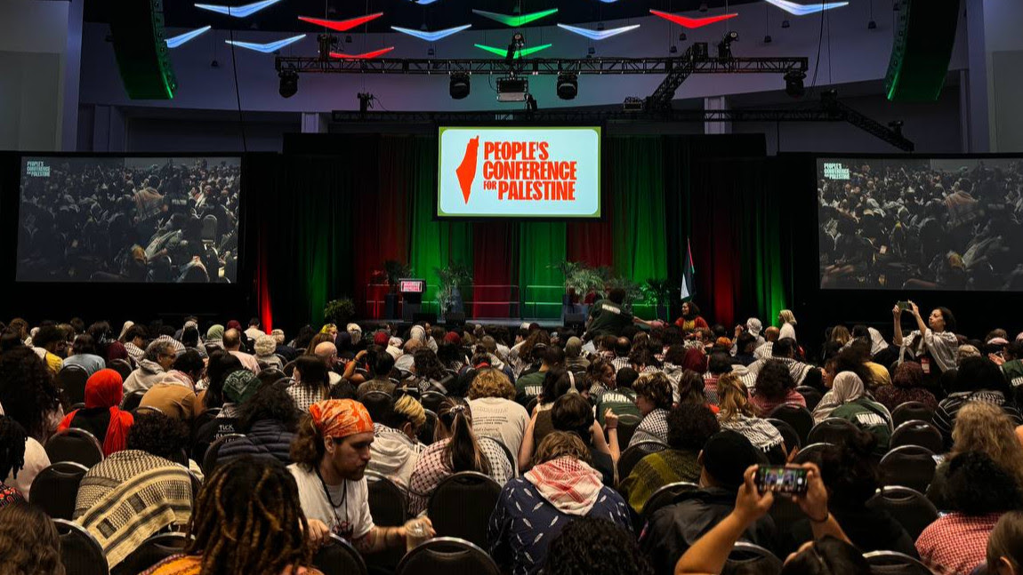 A full conference room, with the middle of three screens on stage with the red logo for the People's Conference for Palestine