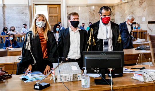 Woman and two men stand in court, some people in background, wearing masks