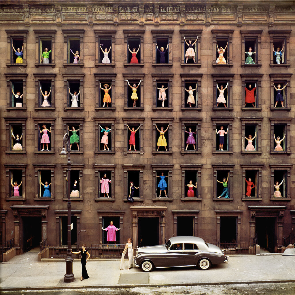 Forty-one women in colored dresses each stand in a window of a building. There is a silver Rolls-Royce on the sidewalk, and two women in gowns are standing near it.