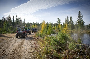 three black, orange and green off-road vehicles going single file down a dirt forest road next to a body of water, forest in background