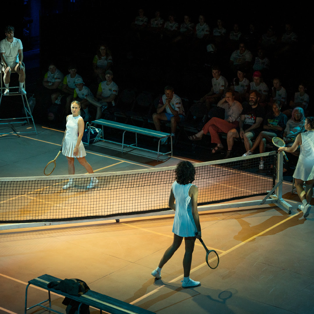 Two women dressed in tennis whites face one another across a tennis net with an audience in the background.
