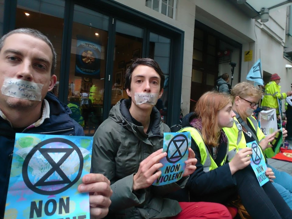 4 rebels sat outside the BBC building. They are all holding blue leaflets which have an extinction symbol on them and the text 'NON-VIOLENT'. They also have tape over their mouths with the words 'silenced by the media' written on.