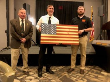 Three people pose for photo holding large wooden American flag