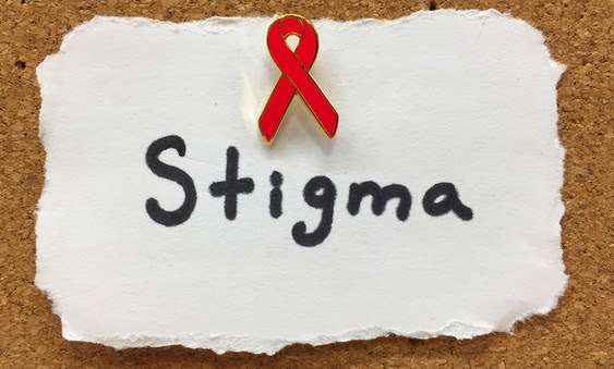 Paper with “stigma” written on it affixed to a corkboard with a pin in the shape of a red ribbon.