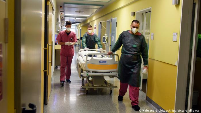 Enfermeiros de máscara empurram cama em corredor de hospital