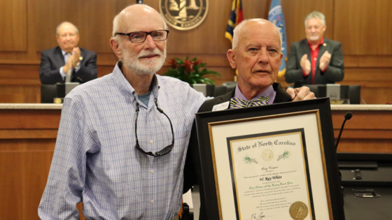 Image of Ray White standing at the Board of Commissioners meeting room podium with Chairman Woodard, who holds a framed certificate.