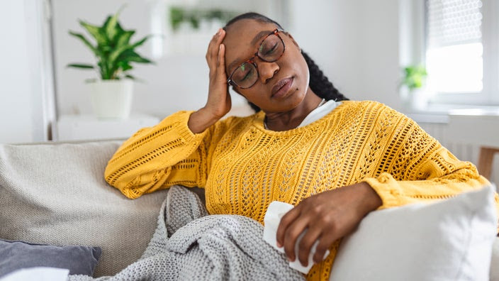 Woman with eyes closed and hand on forehead sitting on the couch with a blanket.