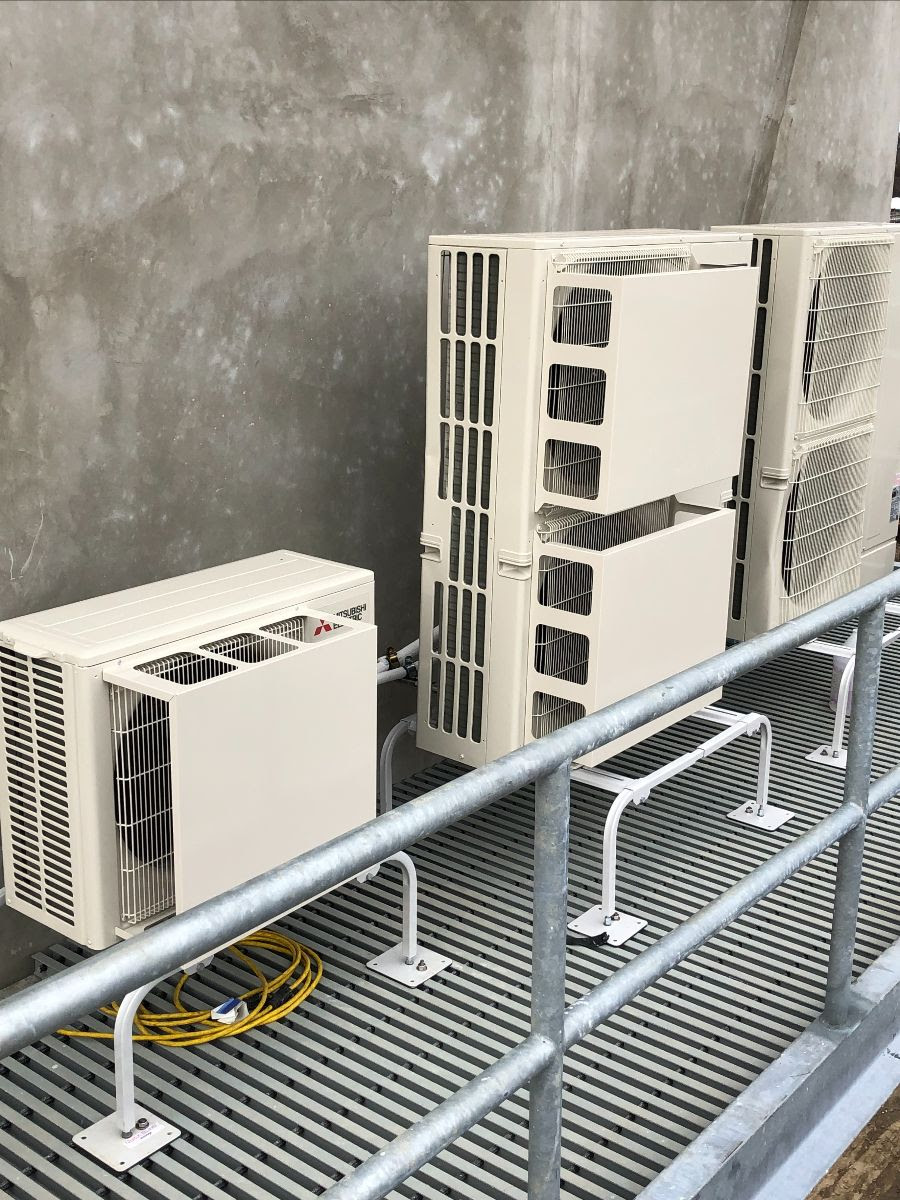 Three HVAC units are shown installed over a metal grate next to a cement wall. The units are bolted to metal legs that lift them off the grate to allow for improved airflow.