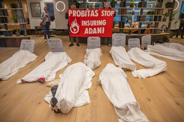 A die-in inside a building. Nine people lie on the floor under white sheets, some with cardboard gravestones at their heads. A red banner in the background reads Probitas stop insuring Adani