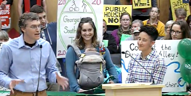 Three people, one holding a baby in a child carrier, speak into microphones at City Hall in front of signs that read 