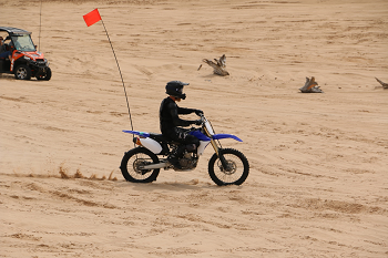 side view of a person wearing a helmet riding a dirt bike over a sandy area, with a side-by-side ORV in the background