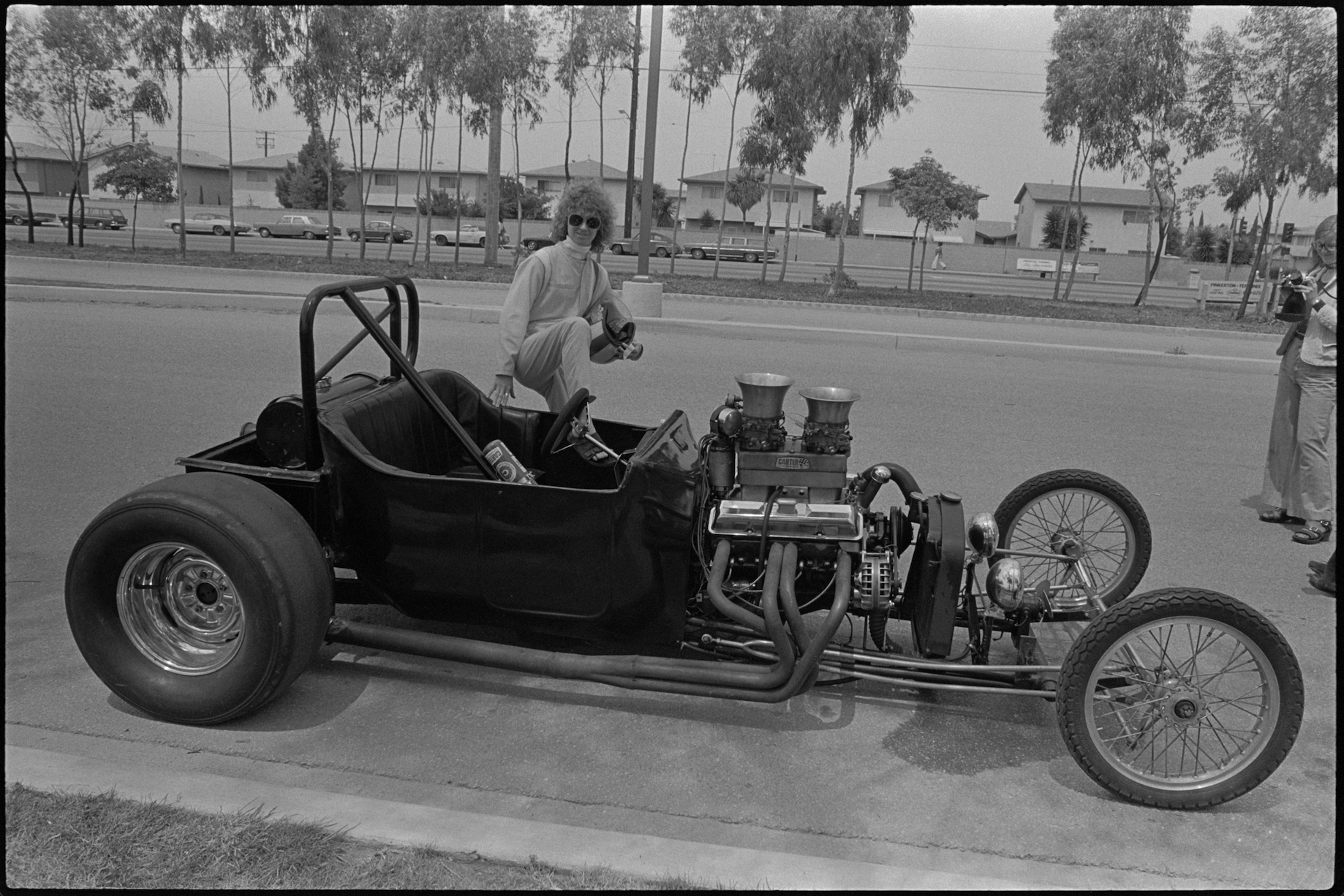 A black-and-white photograph documenting artist Suzanne Lacy's performance "Cinderella in a Dragster" (1976). The artist is stepping into a vintage dragster, wearing sunglasses, a racing jumpsuit, and holding a racing helmet. The vehicle is parked on an empty tree-lined road in California. On the right, a person is taking a photo of Lacy with a small, analog camera.