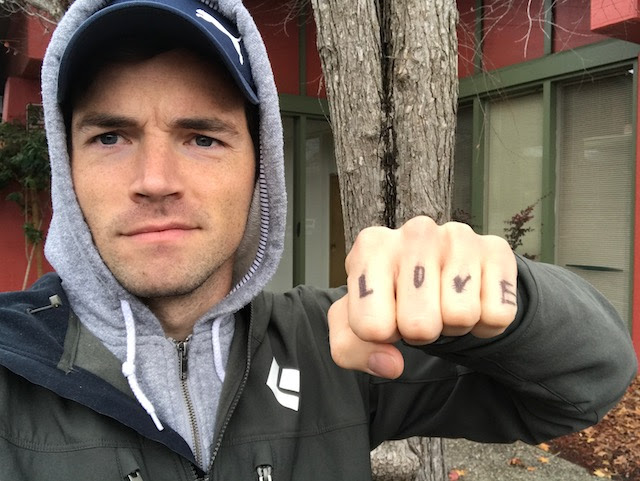 Ian Harding, a man with short brown hair and a black jacket. He is looking serious and lifting his fist with the letters: LOVE.