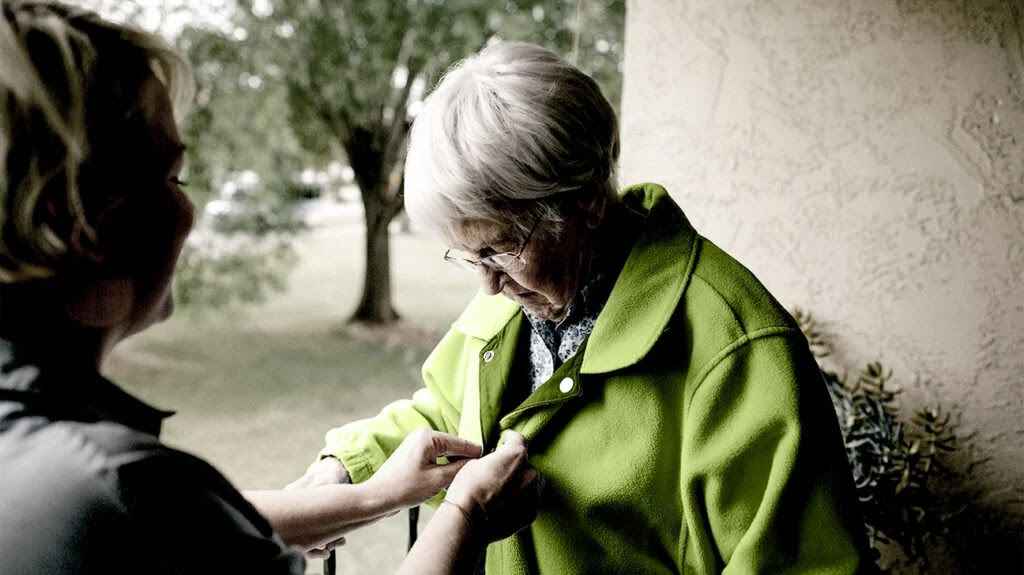 A person helping a woman with Parkinson's disease button up a green coat.