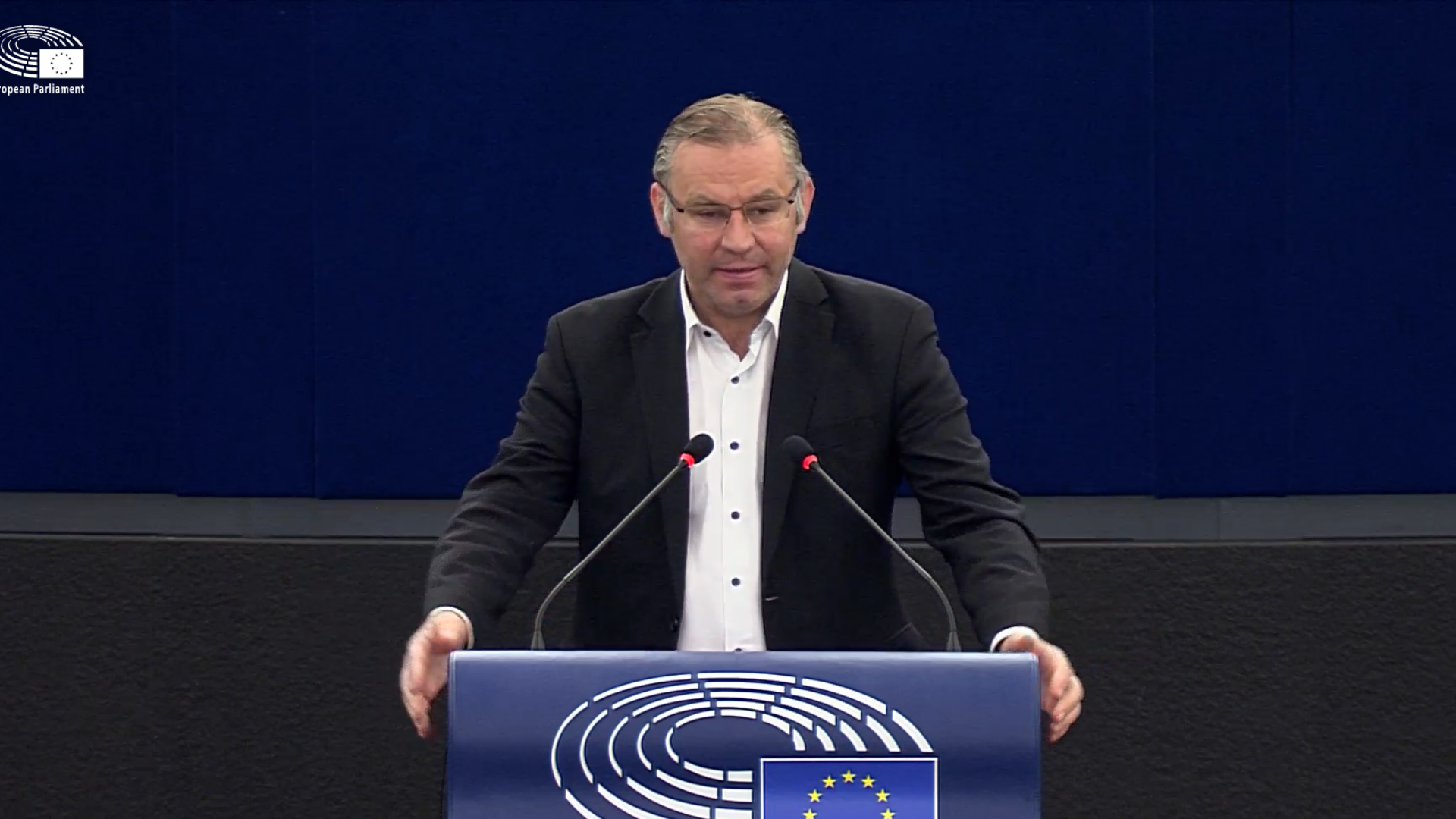 Still from video, grey haired man stands in centre at podium decorated with EU flag, dark blue background