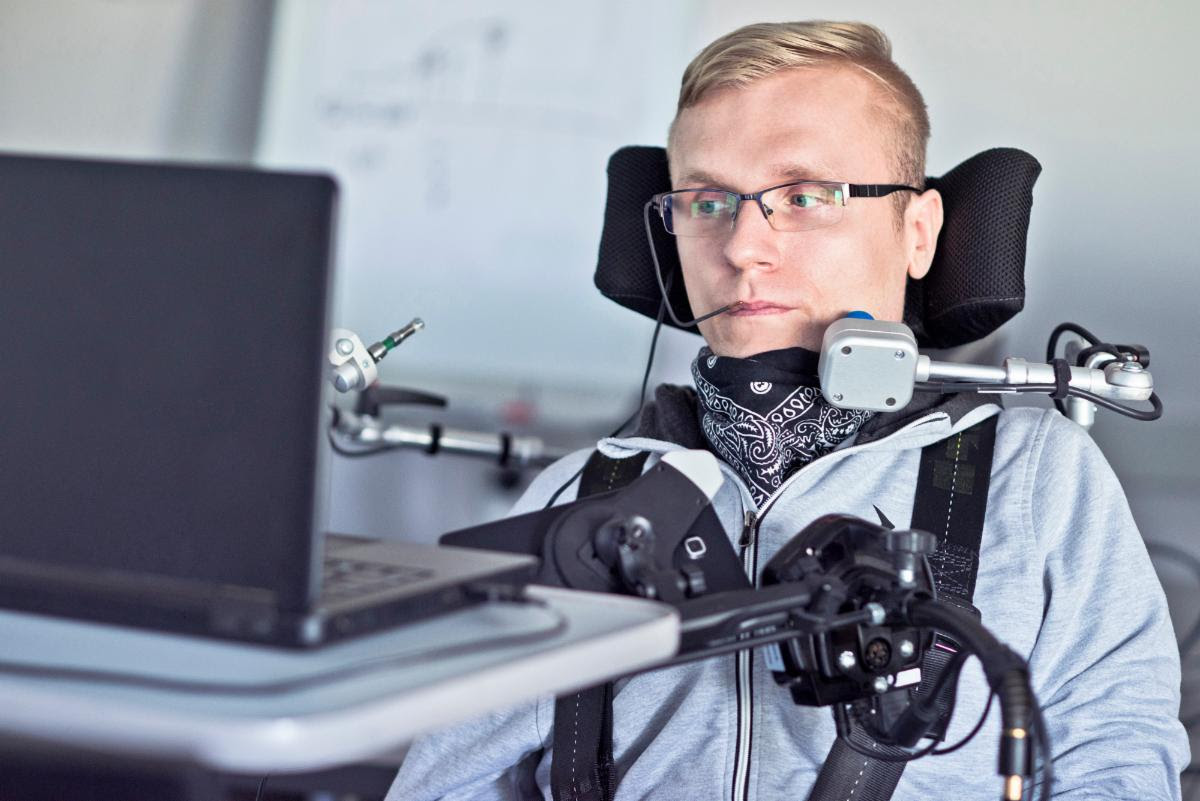 Man with blonde hair and glasses uses a laptop from his wheelchair. He has an adapted computer mouse mounted to his wheelchair .