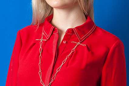  A photograph of a model in a red jumpsuit against a bright blue background. They are wearing a long silver necklace by Sally Cuthbert. 