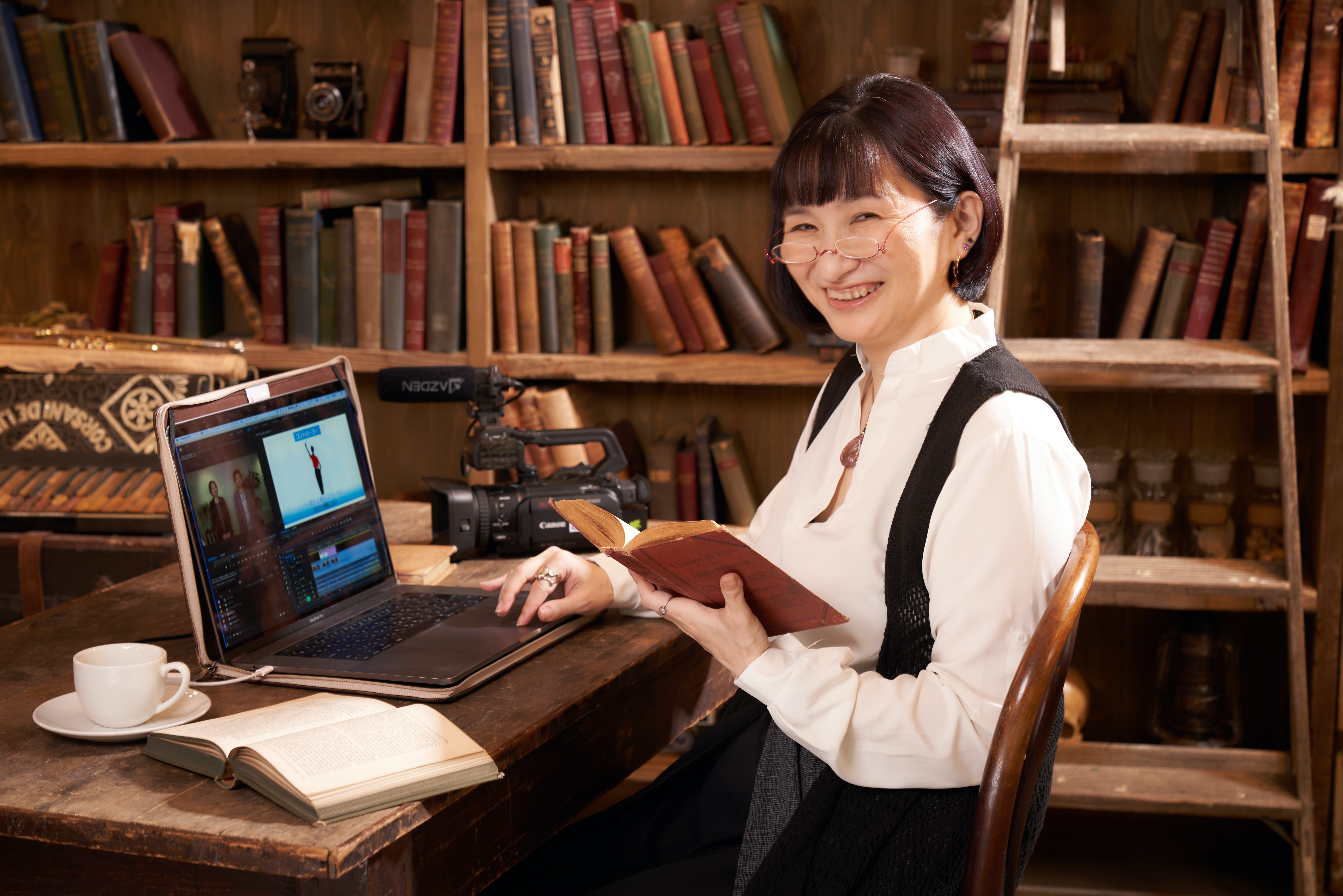 古書の本棚の前でパソコンを開いて動画編集をしながら本を読み微笑む佐藤安南