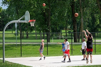 a small group of adult men and young boys wearing athletic shorts, tank tops and T-shirts shoot baskets on a paved basketball court in a greenspace
