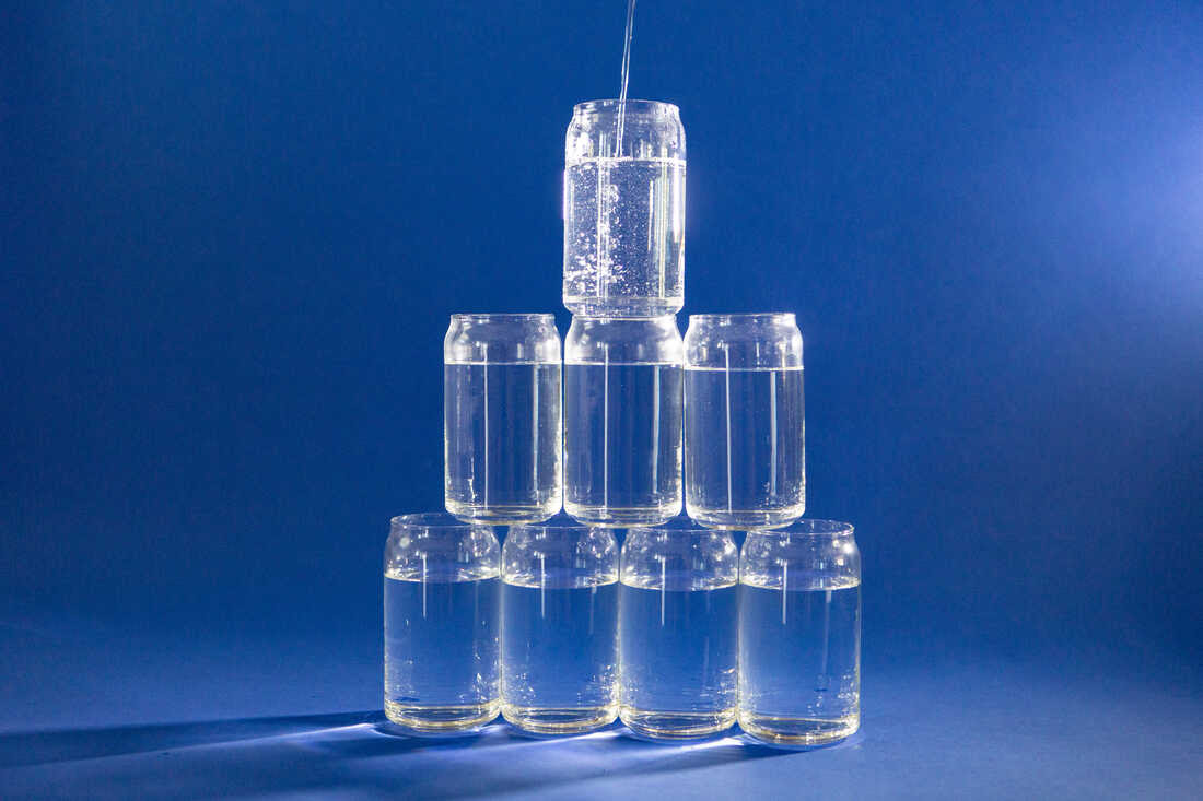 Photograph of eight glasses of water in clear glasses that are stacked into a pyramid. They are against a cobalt blue background and water is being poured into the top glass.