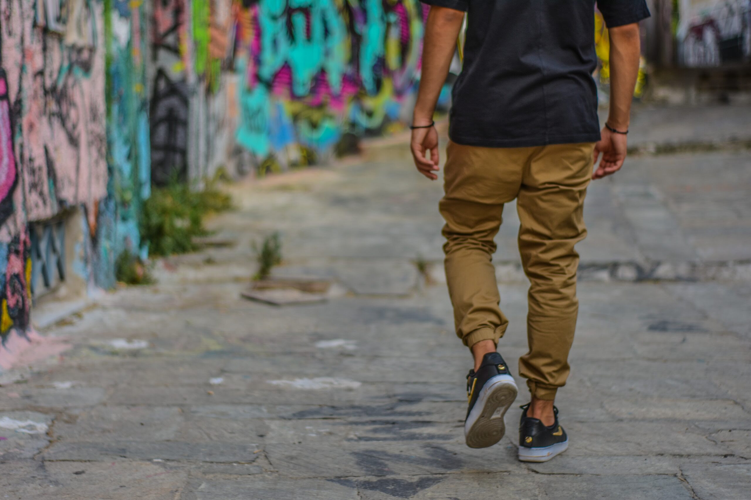 man with sneakers and cuffed pants walks along the road lined with graffiti covered walls