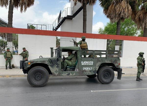 A military vehicle on a road, with one soldier holding a mounted machine gun. Other soldiers are nearby on the street or on the sidewalk. A white wall with a red stripe atop is behind the vehicle.