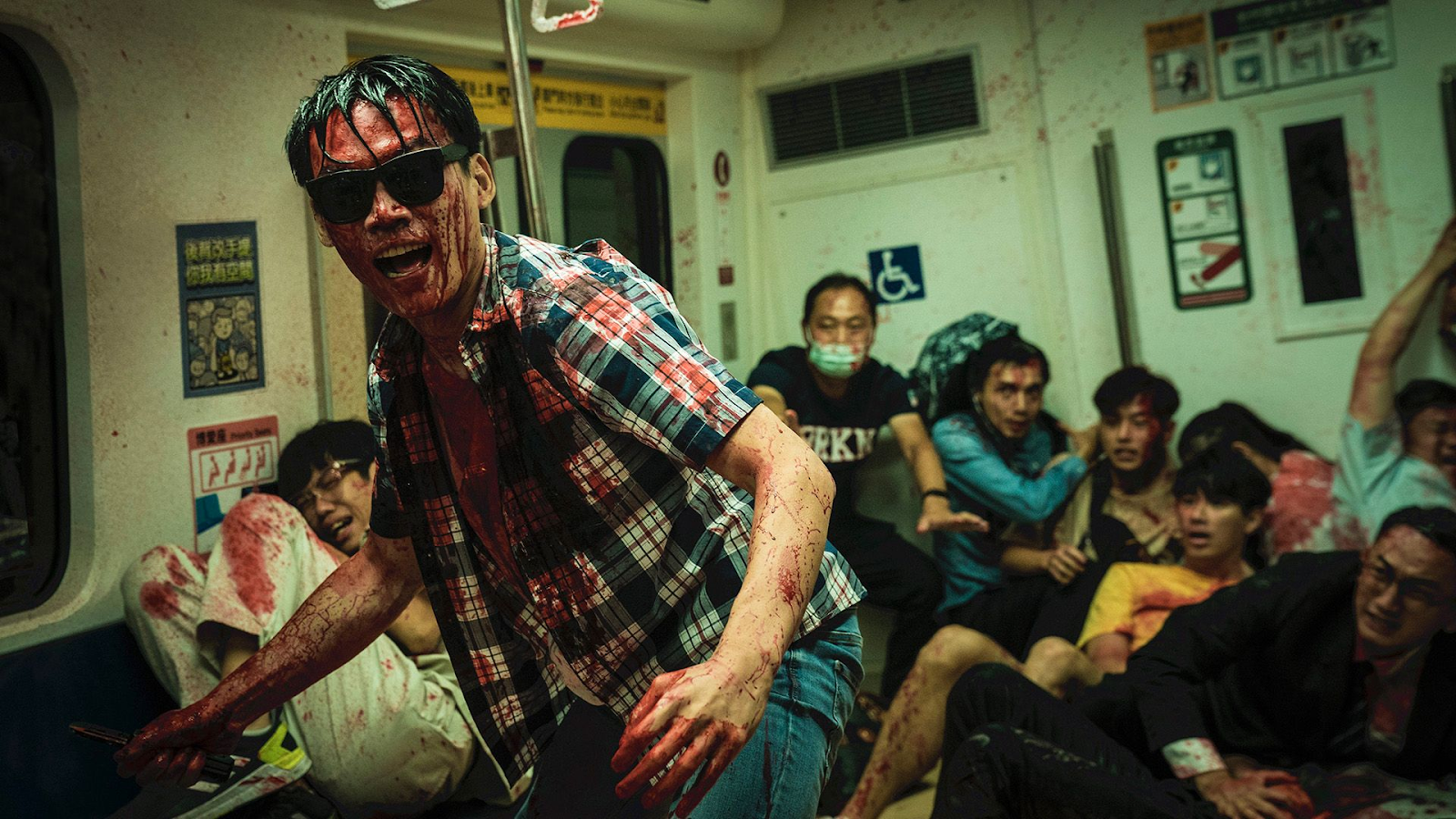 A man drenched in blood wearing sunglasses and a plaid shirt. People are huddled in terror behind him, appearing to be on public transportation.