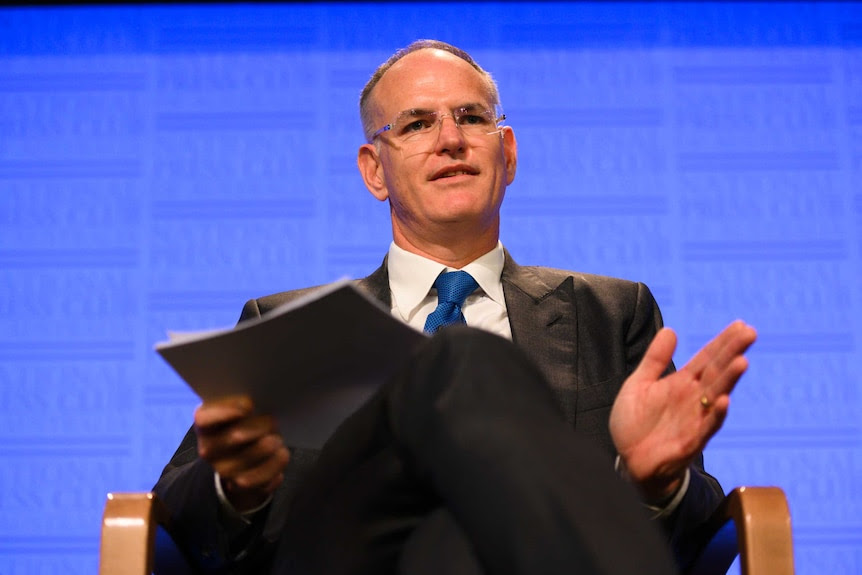 Michael Miller sits on a chair in front of a blue background, wearing a suit.