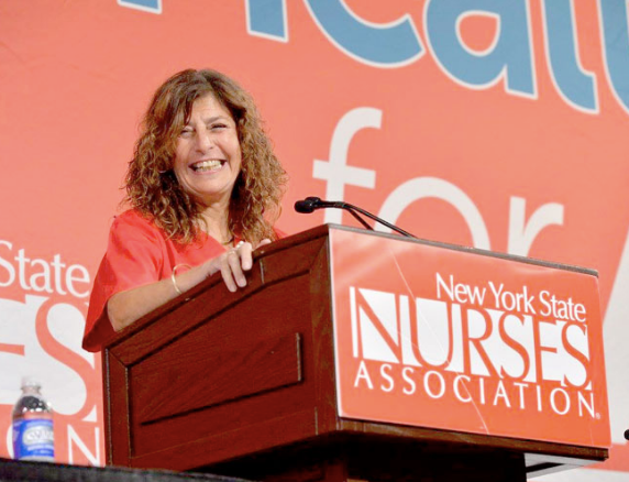 Image of a smiling woman with long curly hair standing at a

podium in a red t-shirt. The front of the podium reads New York State

Nurses Association and the backdrop of the image is a large banner

that says Medicare for All.