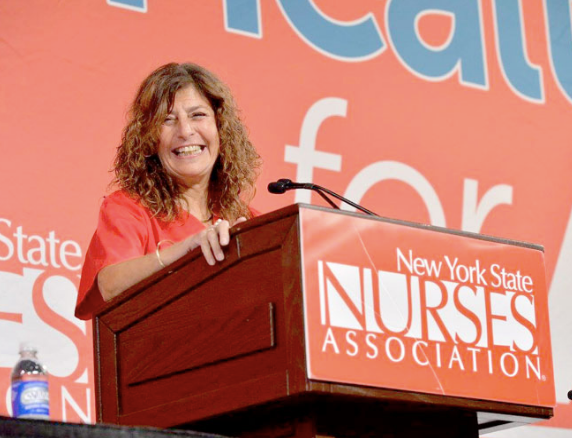 Judy Headshot Image of a smiling woman with long curly hair standing at a
podium in a red t-shirt. The front of the podium reads New York State
Nurses Association and the backdrop of the image is a large banner
that says Medicare for All.