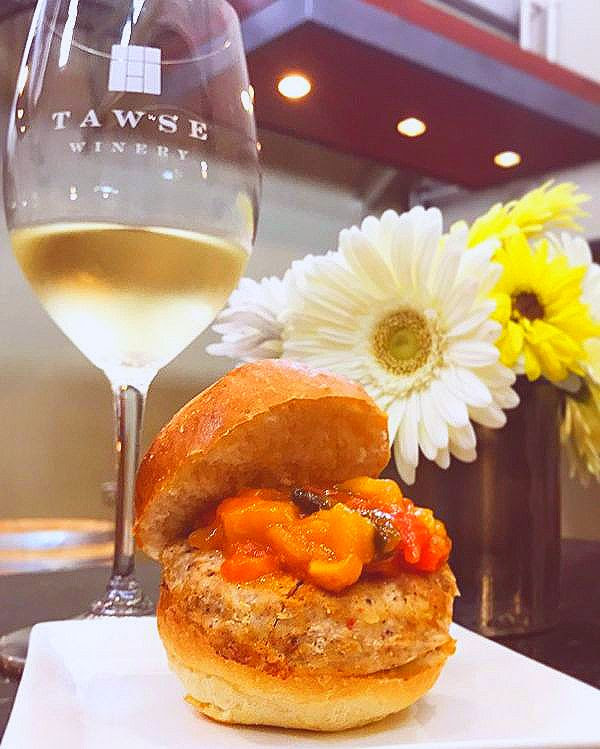 Tawse Winery wine glass containing white wine on a table behind a sumptuous hamburger presentation.