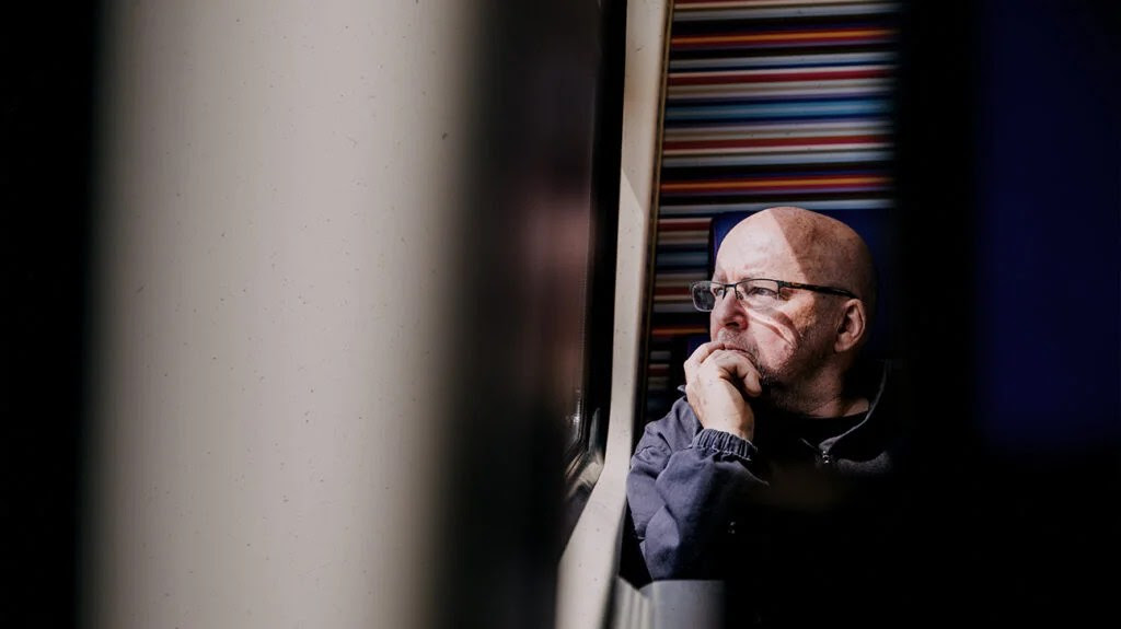 A middle aged man in glasses stares out of a window in deep thought